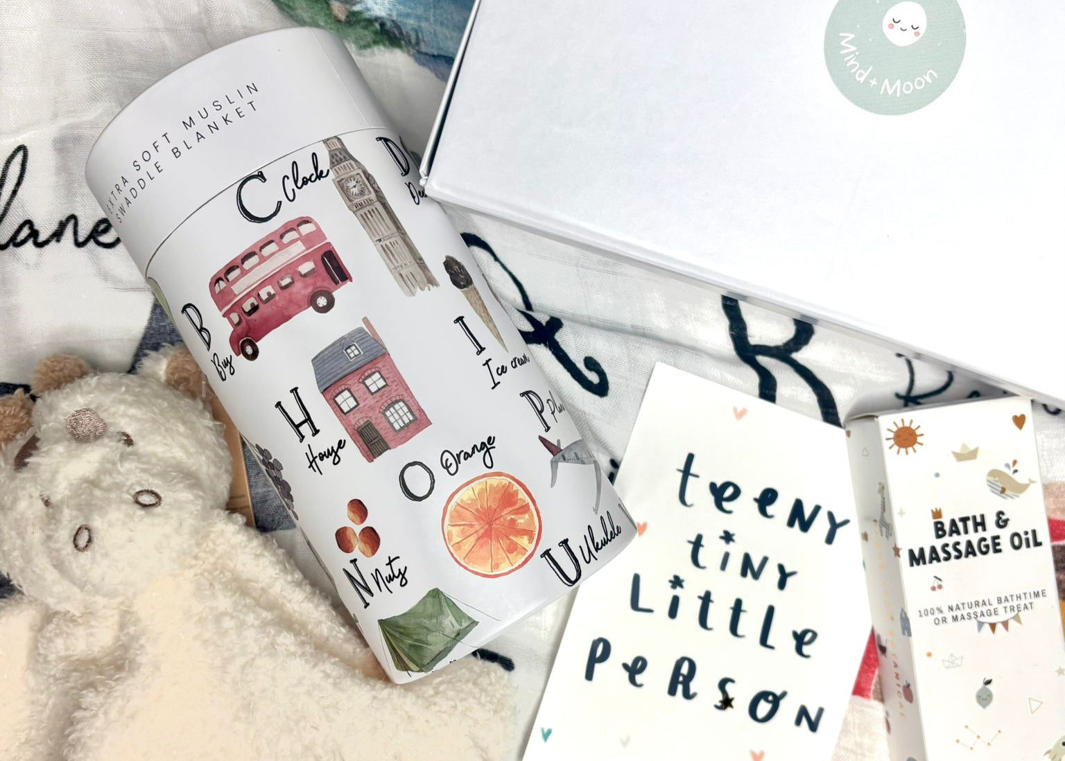 Collection of baby items including a muslin box, cards, and bath products on a soft surface.