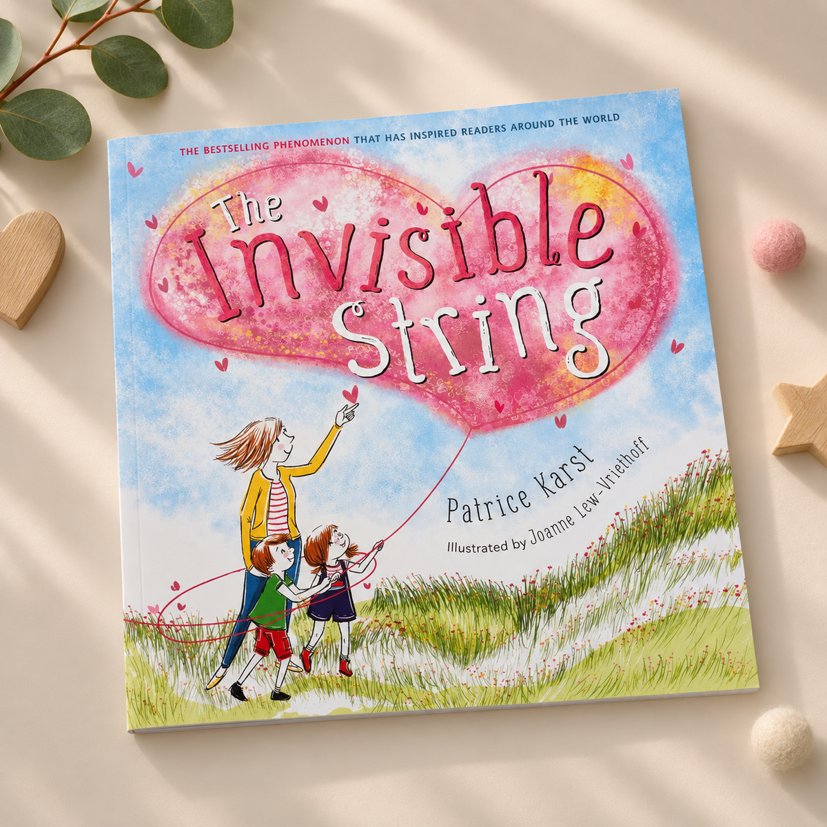 The book cover of 'The Invisible String' by Patrice Karlst and illustrated by Joanna Linzenthal, displayed on a light beige surface with decorative wooden stars, a green leaf, and pom-poms.